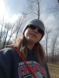 A woman wearing sunglasses and a hat standing in front of trees.