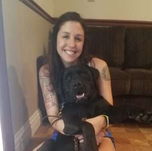 A woman sitting in a chair with her arm around a black dog.